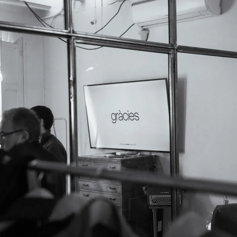 Sala de reunions de l'estudi vista des de fora, a través d'una mampara de vidre i ferro. A la pantalla del fons, la paraula «gràcies».